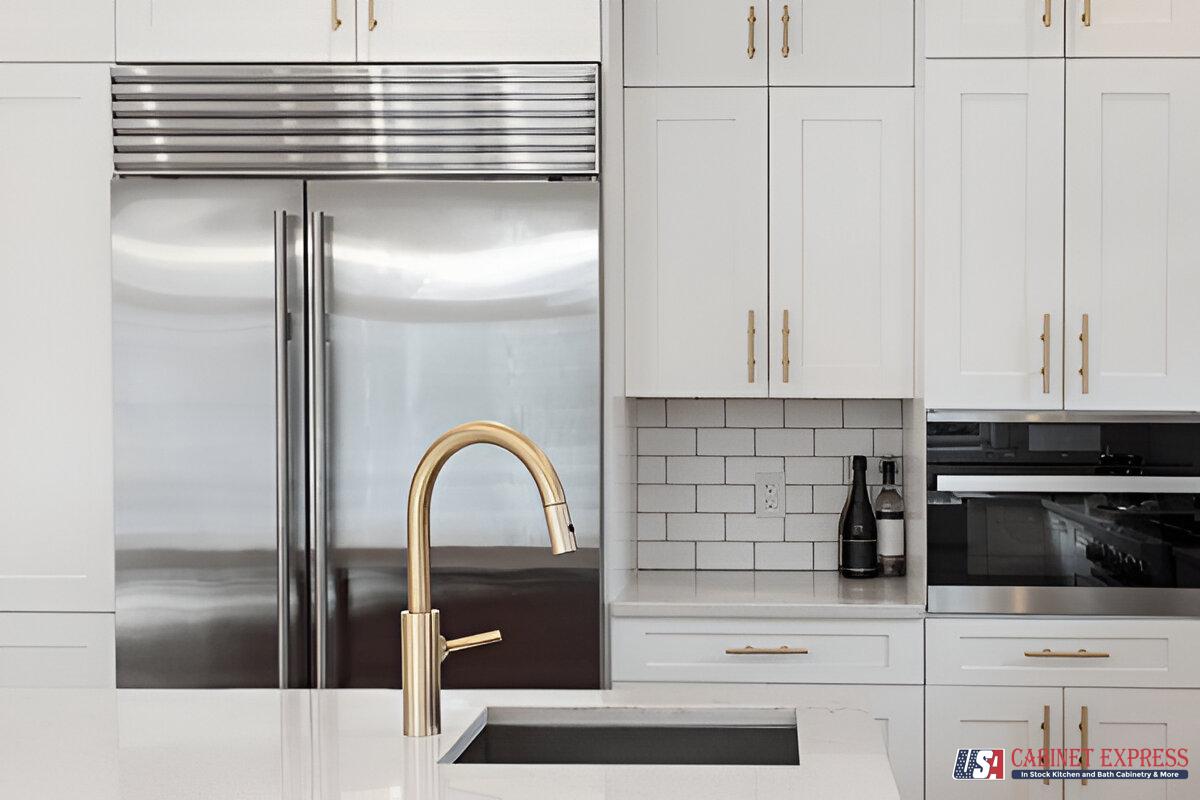 Alt text: "Modern white kitchen featuring a gold faucet and a stainless steel refrigerator. White cabinets with gold handles and a white subway tile backsplash complete the design. The USA Cabinet Express logo is visible in the bottom right corner