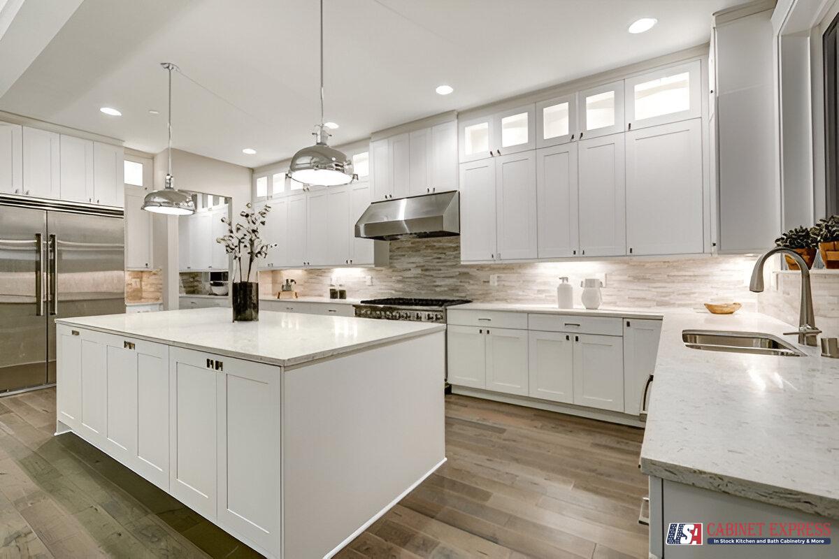 A spacious modern kitchen featuring white shaker cabinets, a large central island with a quartz countertop, and stainless steel appliances. Pendant lights hang above the island, and under-cabinet lighting enhances the neutral-toned backsplash. The USA Cabinet Express logo is visible in the bottom right corner.