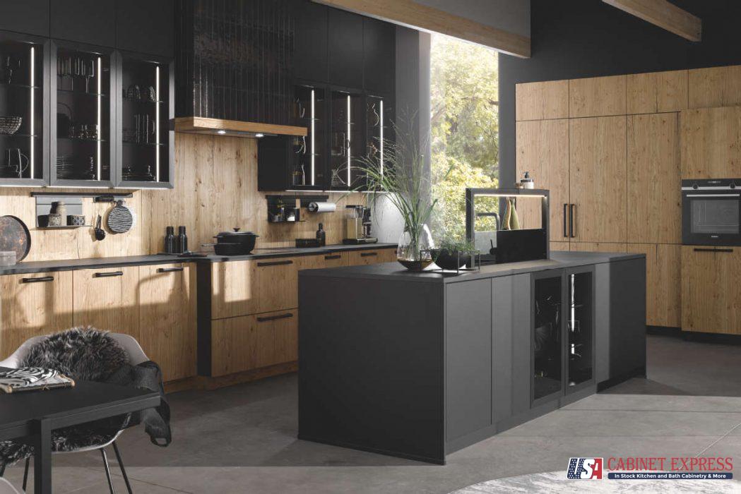 A modern industrial-style kitchen featuring a combination of black and natural wood cabinetry, sleek black countertops, and glass-front upper cabinets. The kitchen includes a spacious black island with integrated storage and a contemporary dining area. Large windows provide natural light and an outdoor view. The USA Cabinet Express logo is visible in the bottom right corner.