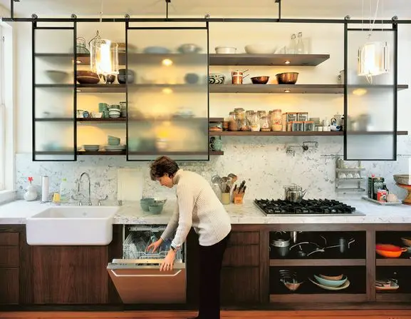How to Organize Your Kitchen Cabinets Like a Pro (Step-by-Step Guide) 6 A modern kitchen with sliding frosted glass cabinets, open wooden shelves, a farmhouse sink, and a marble backsplash, featuring a woman loading a dishwasher.