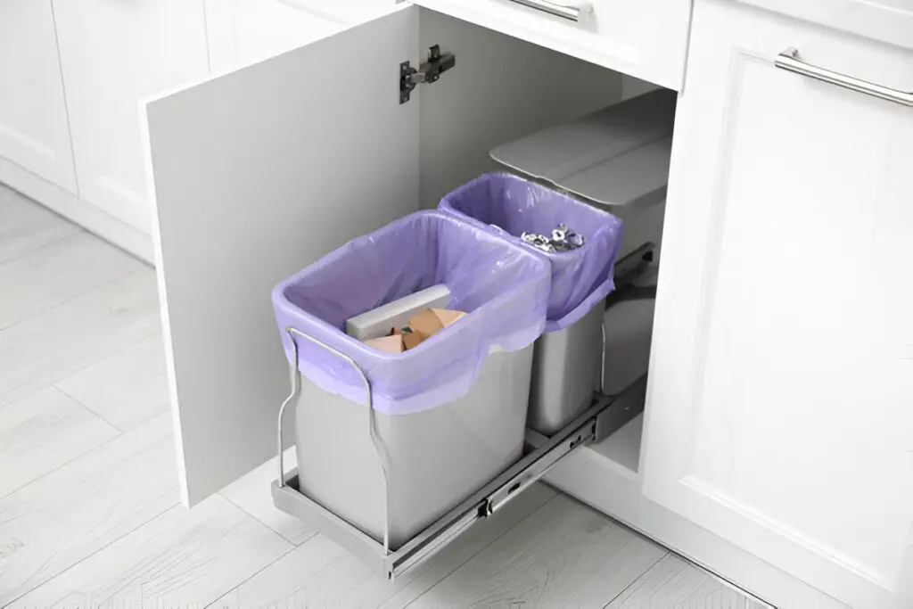 How to Organize Your Kitchen Cabinets Like a Pro (Step-by-Step Guide) 14 A pull-out kitchen trash and recycling bin system inside a white cabinet, featuring two separate bins lined with purple trash bags.