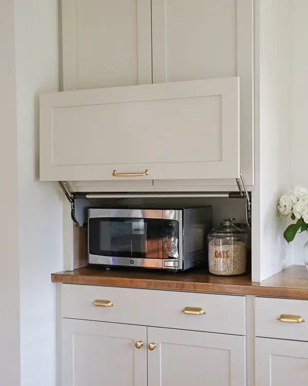 How to Organize Your Kitchen Cabinets Like a Pro (Step-by-Step Guide) 16 A modern kitchen appliance garage with a lift-up cabinet door, concealing a microwave and storage space on a wooden countertop with gold hardware.
