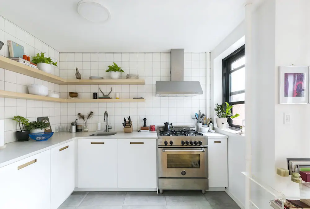 Drawer vs. Door Cabinets: Which Kitchen Storage Style Works Best? 4 Modern minimalist kitchen with white cabinets, open wooden shelves, a stainless steel stove, and a tiled backsplash, featuring greenery and stylish decor.
