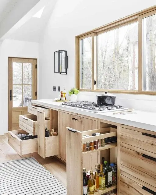 A modern kitchen with natural wood cabinetry, large windows, and pull-out storage solutions for spices, utensils, and cookware.