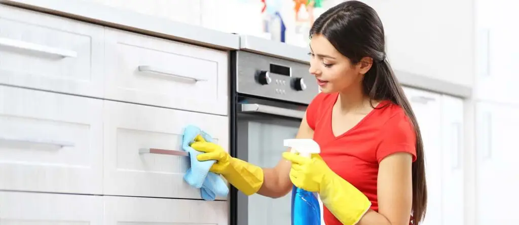 How to Organize Your Kitchen Cabinets Like a Pro (Step-by-Step Guide) 18 A woman wearing yellow rubber gloves and a red shirt cleaning white kitchen cabinets with a blue cloth and a spray bottle.