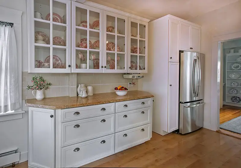 A bright and elegant kitchen with white cabinetry, glass-front upper cabinets displaying decorative dishware, a beige granite countertop, and a stainless-steel refrigerator.