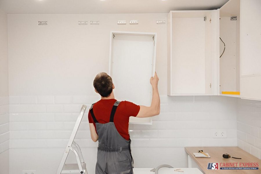 A worker wearing a red shirt and gray overalls installing a white kitchen cabinet on a tiled wall. A step ladder and various tools are visible in the background. The USA Cabinet Express logo is displayed in the bottom right corner.